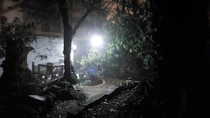 Un espace extérieur peu éclairé, avec une table et des chaises, entouré d’arbres et de feuillage.