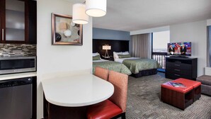A hotel room with a kitchenette, a dining area, two beds, a TV, and a balcony.