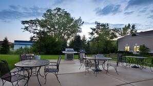 Une terrasse extérieure avec des tables et des chaises en métal, un barbecue et un grand arbre en arrière-plan.