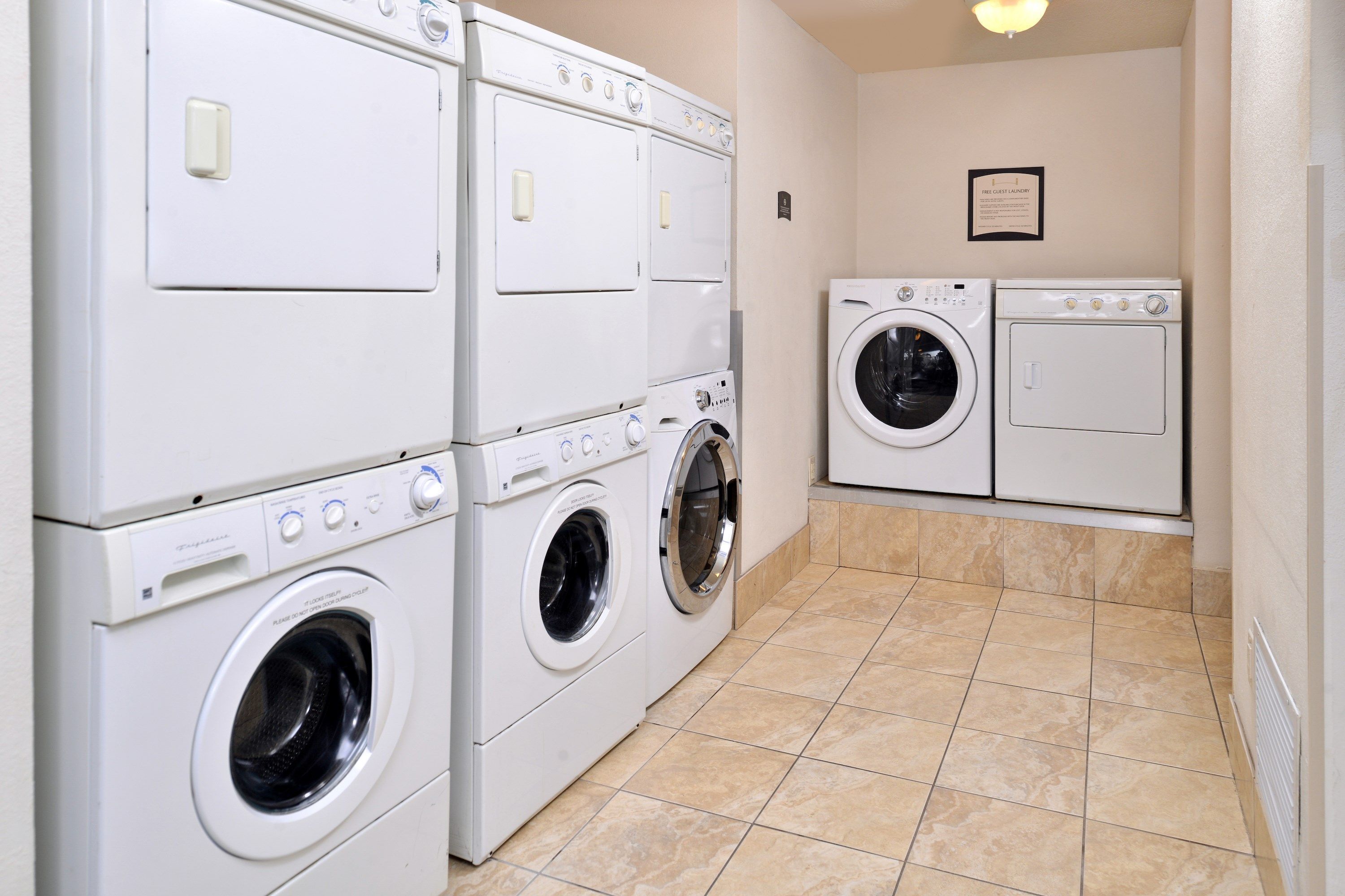 A laundry room with stacked washers and dryers, a tiled floor, and a wall-mounted sign.
