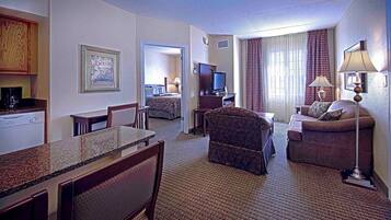A hotel room with a dining area, a living room with a sofa and armchair, a TV, and a kitchenette.