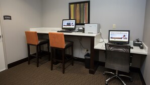 Un espace de travail comprenant deux bureaux, chacun équipé d’un écran d’ordinateur, d’un clavier et d’une souris. Il y a deux chaises, dont une avec un coussin et une autre avec un dossier en tissu ajouré. Une poubelle se trouve sous les bureaux, et un tableau encadré est accroché au mur.