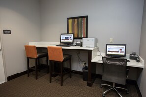 Un espace de travail comprenant deux bureaux, chacun équipé d’un écran d’ordinateur, d’un clavier et d’une souris. Il y a deux chaises, dont une avec un coussin et une autre avec un dossier en tissu ajouré. Une poubelle se trouve sous les bureaux, et un tableau encadré est accroché au mur.