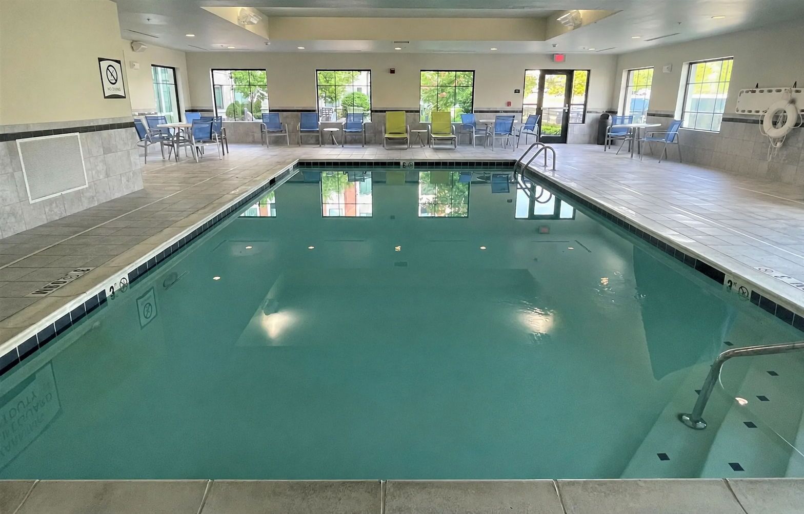 An indoor swimming pool with clear water, surrounded by blue chairs and tables, and large windows allowing natural light.