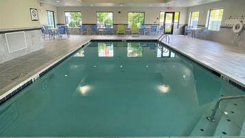 An indoor swimming pool with clear water, surrounded by blue chairs and tables, and large windows allowing natural light.
