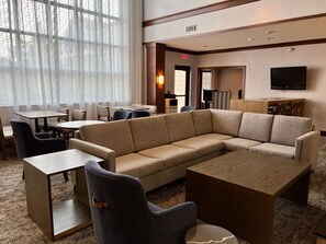 A hotel lobby with a sectional sofa, round tables, and a flat-screen TV mounted on the wall.