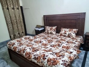 A bed with a floral bedspread and two pillows, a wooden headboard, a nightstand with a lamp, and a curtain with a pattern. - Remarkable 3-bedroom (Gujar Khan)
