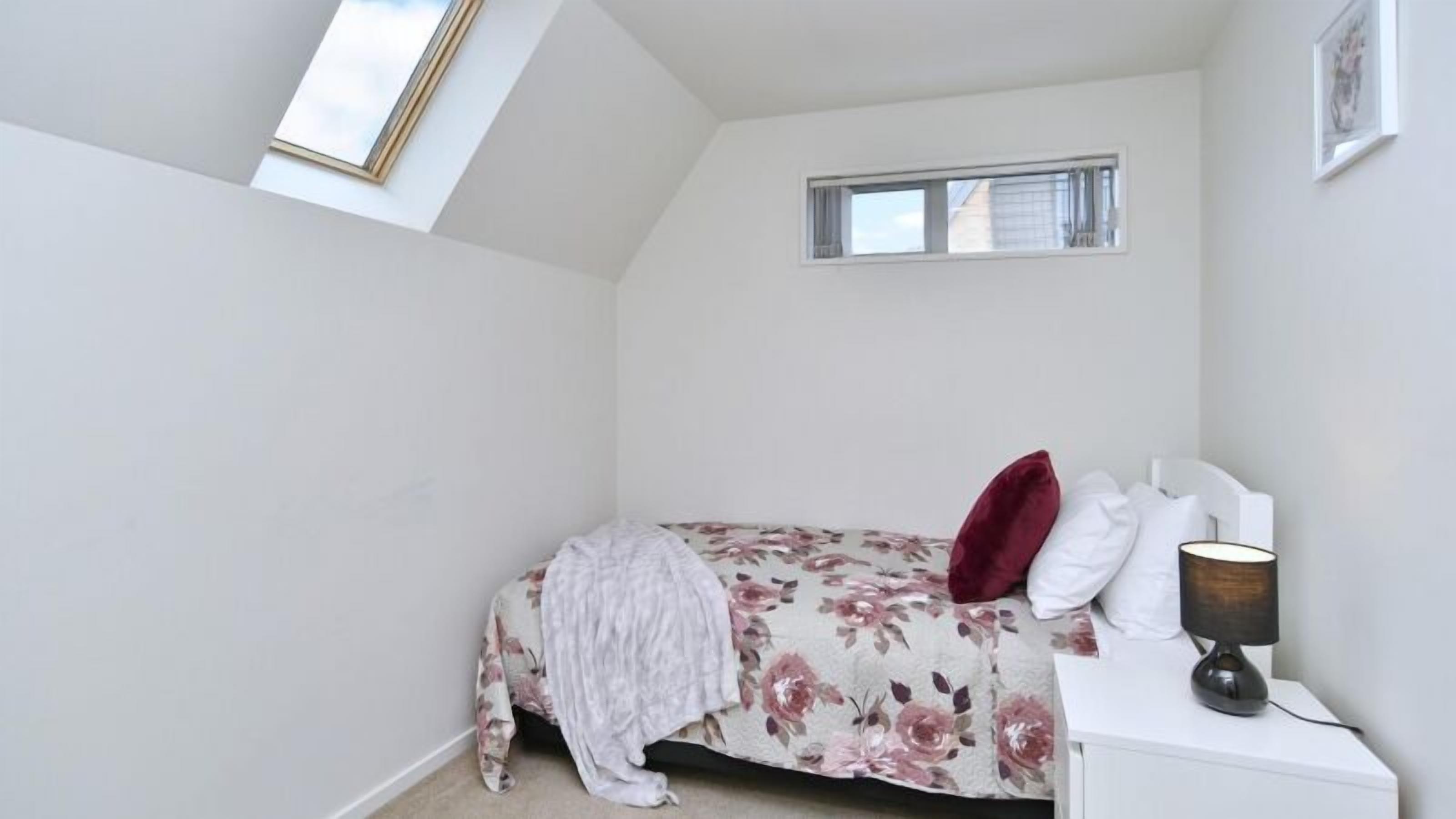 A bedroom with a sloped ceiling, a skylight, a bed with floral bedding, a bedside table with a lamp, and a window with curtains.