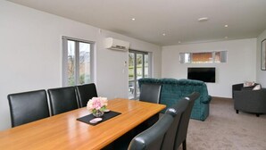 A modern living room with a wooden dining table, black chairs, a teal sofa, and a flat-screen TV mounted on the wall.