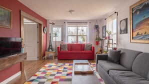 A living room with a red sofa, a grey sofa, a TV, and a colorful rug. - Artistic Getaway Near Halifax | King Bed, Spacious Deck & Fire Pit (Upper Hammonds Plains)
