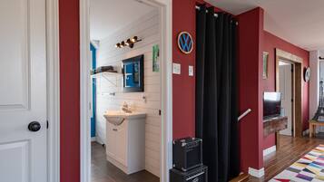 Um corredor com parede vermelha, porta branca, banheiro com pia e espelho, e uma TV em uma estante de madeira.