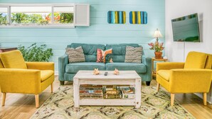 A living room with a teal sofa, two yellow armchairs, a coffee table with books, a TV, and a window with blinds. - Charming 3 Bedroom Suite 200 steps to Beach, car available (Freeport)