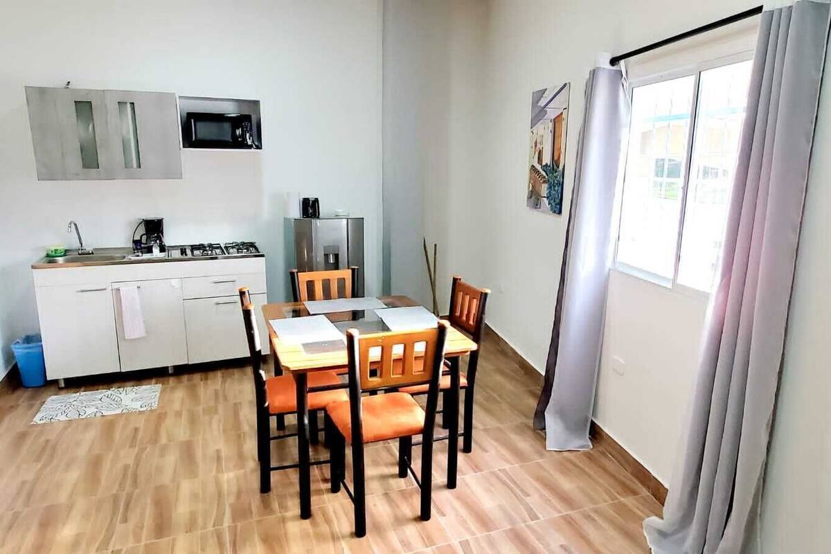 A compact kitchen with a microwave, stove, and refrigerator, a dining table with chairs, and a window with curtains.