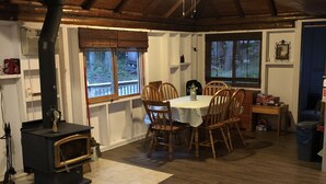 A rustic dining area with a wooden table and chairs, a black wood stove, and a window overlooking a wooden deck. - Lake Front Cottage Property  (Bracebridge)