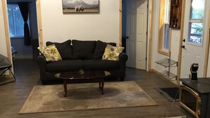 A living room with a dark-colored sofa, a coffee table, and a wooden floor. There is a door with a window on the right side, and a picture on the wall above the sofa. - Lake Front Cottage Property  (Bracebridge)