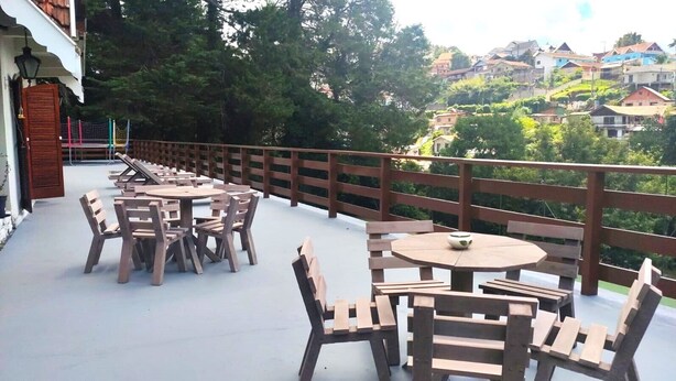 Een terras buiten met houten tafels en stoelen, een houten omheining en uitzicht op een heuvel met huizen.