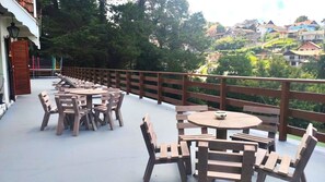 Eine Terrasse im Freien mit Holztischen und -stühlen, einem Holzzaun und Blick auf eine Hügelkuppe mit Häusern.