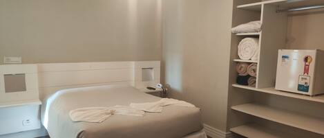 Quarto de hotel com cama, mesa de cabeceira, aquecedor fixado na parede e estante com toalhas e um aquecedor.