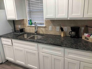 A kitchen with white cabinets, a granite countertop, a stainless steel sink, a window with blinds, and a coffee maker. - 3 bedroom tiny home (Wichita Falls)