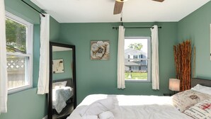 Ein Schlafzimmer mit Bett, Deckenventilator, Fenster mit Vorhängen, Spiegel und Wandlampe.
