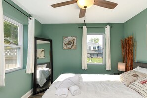 Ein Schlafzimmer mit Bett, Deckenventilator, Fenster mit Vorhängen, Spiegel und Wandlampe.