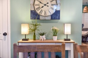 A room with a wall-mounted clock, two lamps, and two potted plants on a wooden console table.