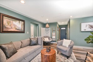 Un salon avec un canapé gris, une table basse ronde et un fauteuil. Il y a un tableau au mur et une porte à l’arrière-plan.