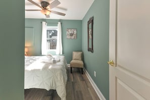 A bedroom with a bed, a chair, a ceiling fan, a window with curtains, and a door.