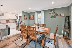 A modern kitchen with white cabinets, a dining area with wooden chairs and a table, and a staircase with a glass railing.