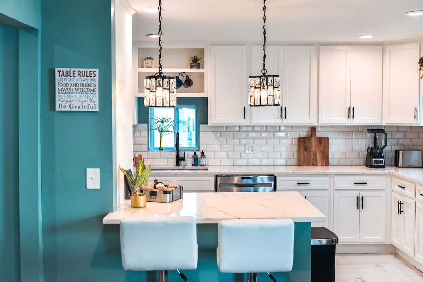 Une cuisine moderne avec des meubles blancs, un îlot central, des luminaires suspendus et un panneau indiquant les règles à respecter à table.