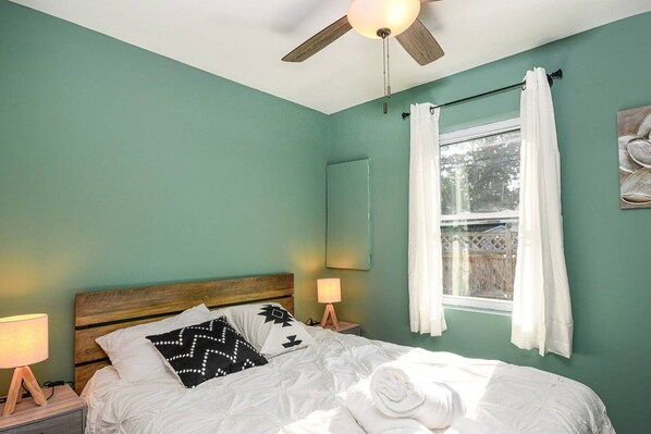 A bedroom with a bed, a ceiling fan, a window with curtains, and a nightstand with a lamp.