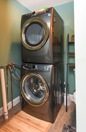 A stacked washer and dryer unit with a control panel, hoses, and a wooden floor.