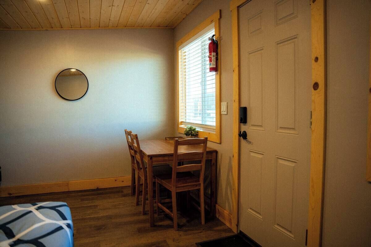 A room with a wooden dining table and chairs, a round mirror on the wall, a window with blinds, and a white door.