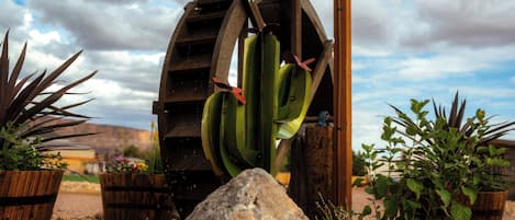 Une roue à eau ornée de bananes vertes suspendues, entourée de barils en bois et de plantes en pot.