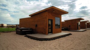 Una cabaña de madera moderna con puerta de cristal, ubicada en una zona arenosa bajo un cielo despejado.