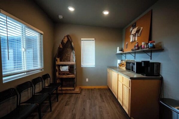 A kitchen with wooden cabinets, a microwave, and a coffee maker. There are two windows with blinds, a wooden bench, and a bulletin board on the wall. - Desert Sage Retreat (Colorado City)