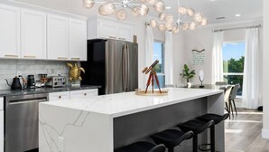 A modern kitchen with white cabinetry, a large island with bar stools, a stainless steel refrigerator, and a unique chandelier. - Lakefront Home. Insane Views - Pool/Spa! Gameroom! (Kingsland)