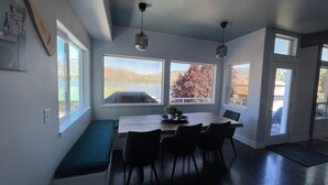 Un coin repas avec une table en bois, des chaises et un banc. De grandes fenêtres offrent une vue sur un lac et des arbres.