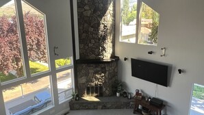Un vaste salon doté d’une cheminée en pierre, de grandes fenêtres et d’un téléviseur à écran plat fixé au mur.