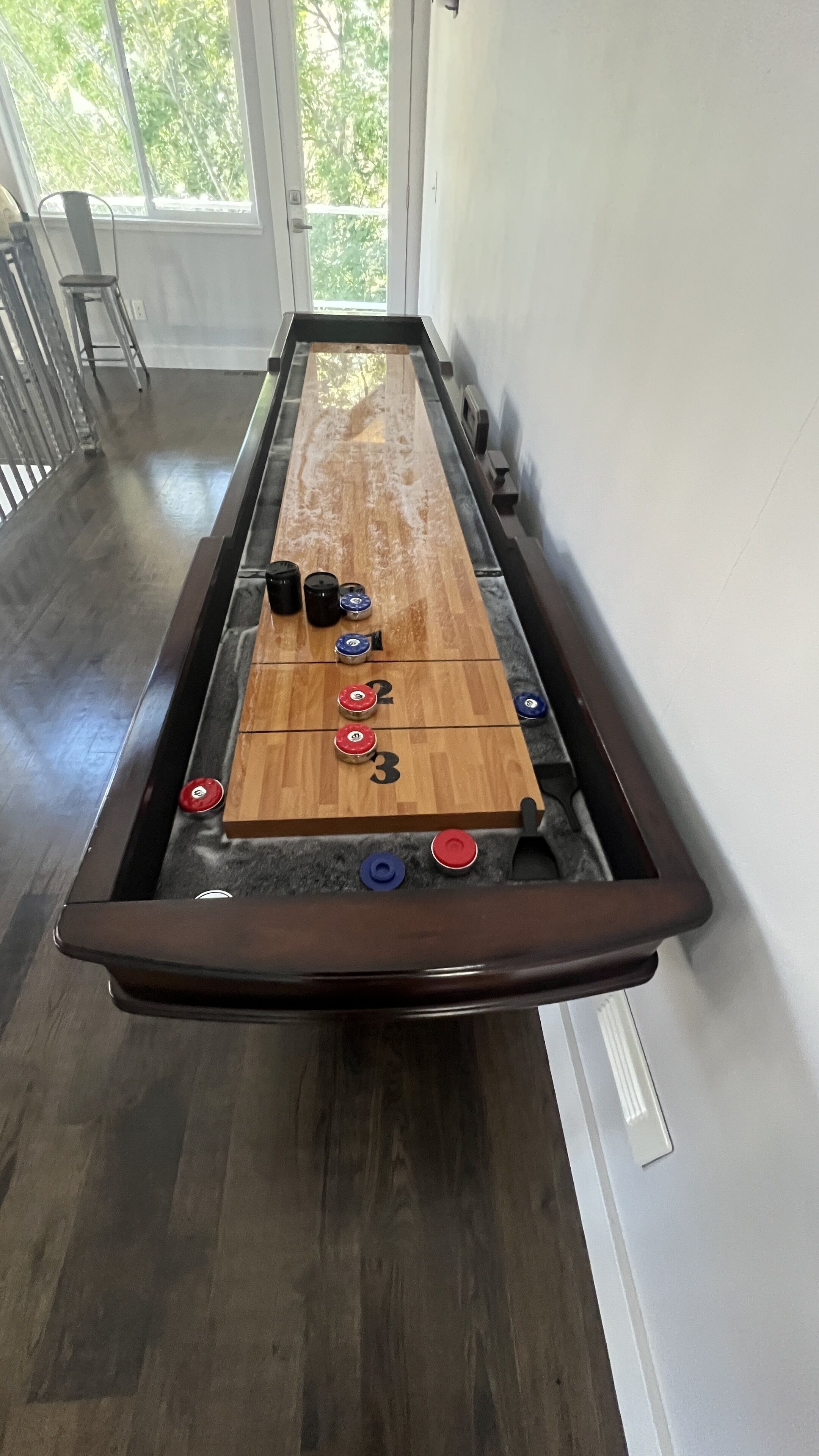 A shuffleboard table with numbered scoring areas and pucks, set against a white wall with a window.