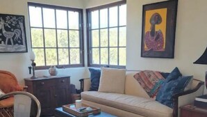 Un salón con sofá, reposapiés y una alfombra estampada. Se ven ventanas con vistas a árboles, un cuadro en la pared y una lámpara sobre una mesita.