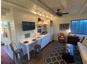 Un espacio habitable compacto con kitchenette, comedor y zona de estar con sofá y sillones.