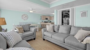A living room with a grey sofa, a coffee table, and a TV. There is a kitchen area with cabinets and a microwave. - "Island Time", Peaceful 5BR Oceanfront Cottage (Surf City)