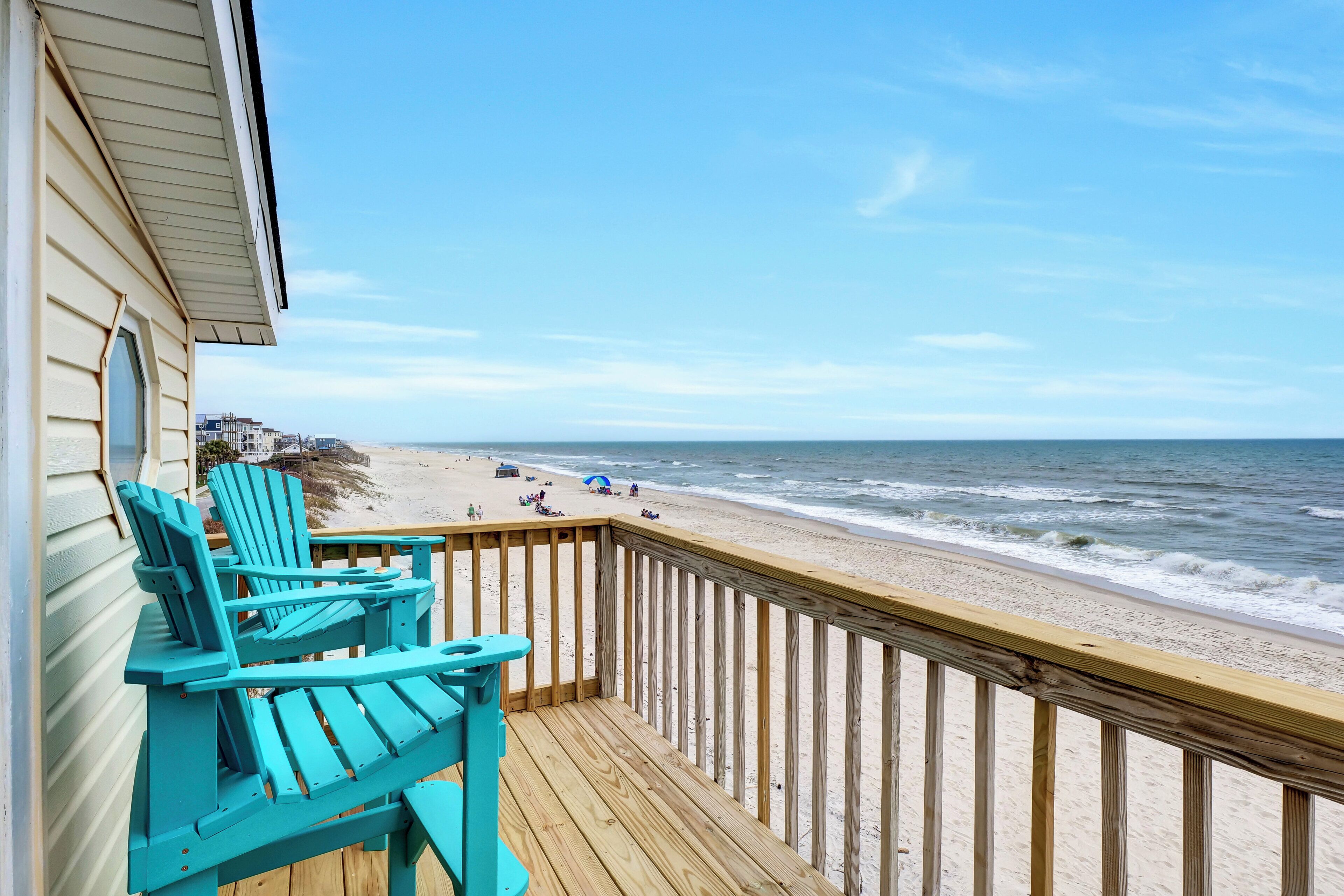 A wooden deck with turquoise Adirondack chairs overlooking a beach with people and a boardwalk.