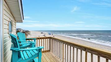 A wooden deck with turquoise Adirondack chairs overlooking a beach with people and a boardwalk.