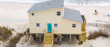 A beach house with a staircase leading to the entrance, situated on a sandy beach with the ocean in the background.