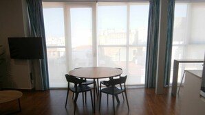 A dining area with a round table and four chairs, a flat-screen TV mounted on the wall, and a view of buildings through large windows.