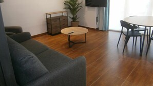A living room with a sofa, a coffee table, a TV, a dining table with chairs, a plant, and a cabinet.