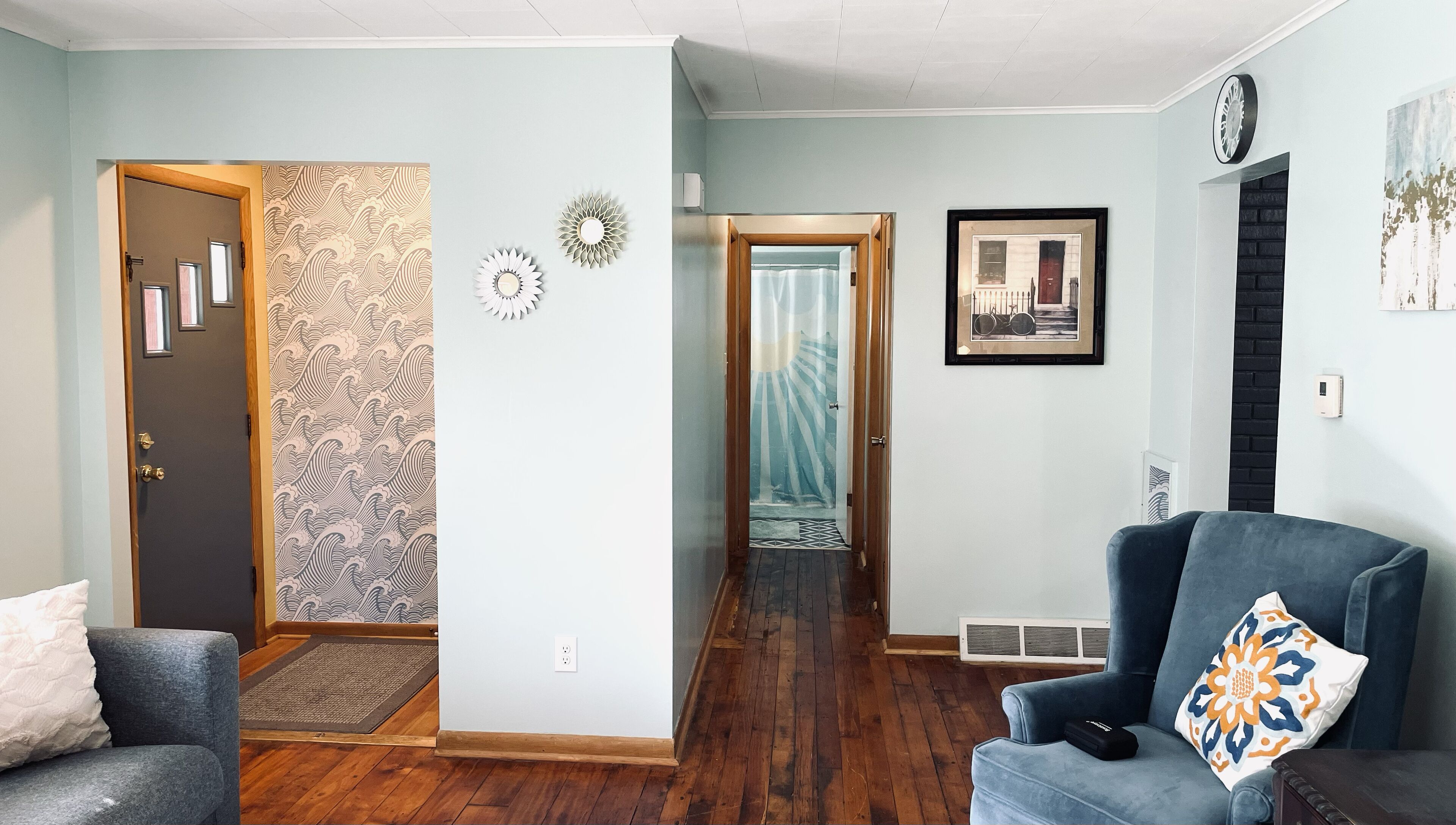 A living room with a blue armchair, a wooden door with a patterned wallpaper, and a hallway leading to another room.