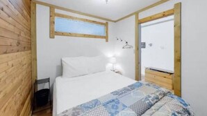 A bedroom with a wooden wall, a bed with white bedding, a bedside table with a lamp, and a window above the bed. - Lakefront Suite w/ Private Hot Tub | Fireplace (Naramata)